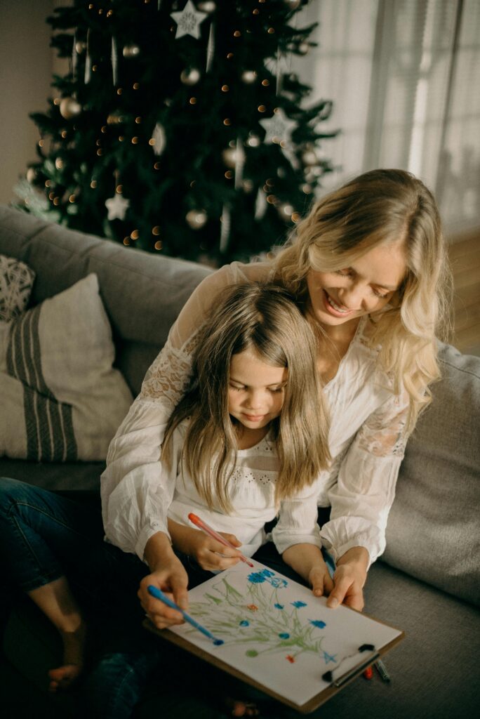 A mother and daughter enjoy drawing together by a Christmas tree, capturing a cozy holiday moment indoors.
