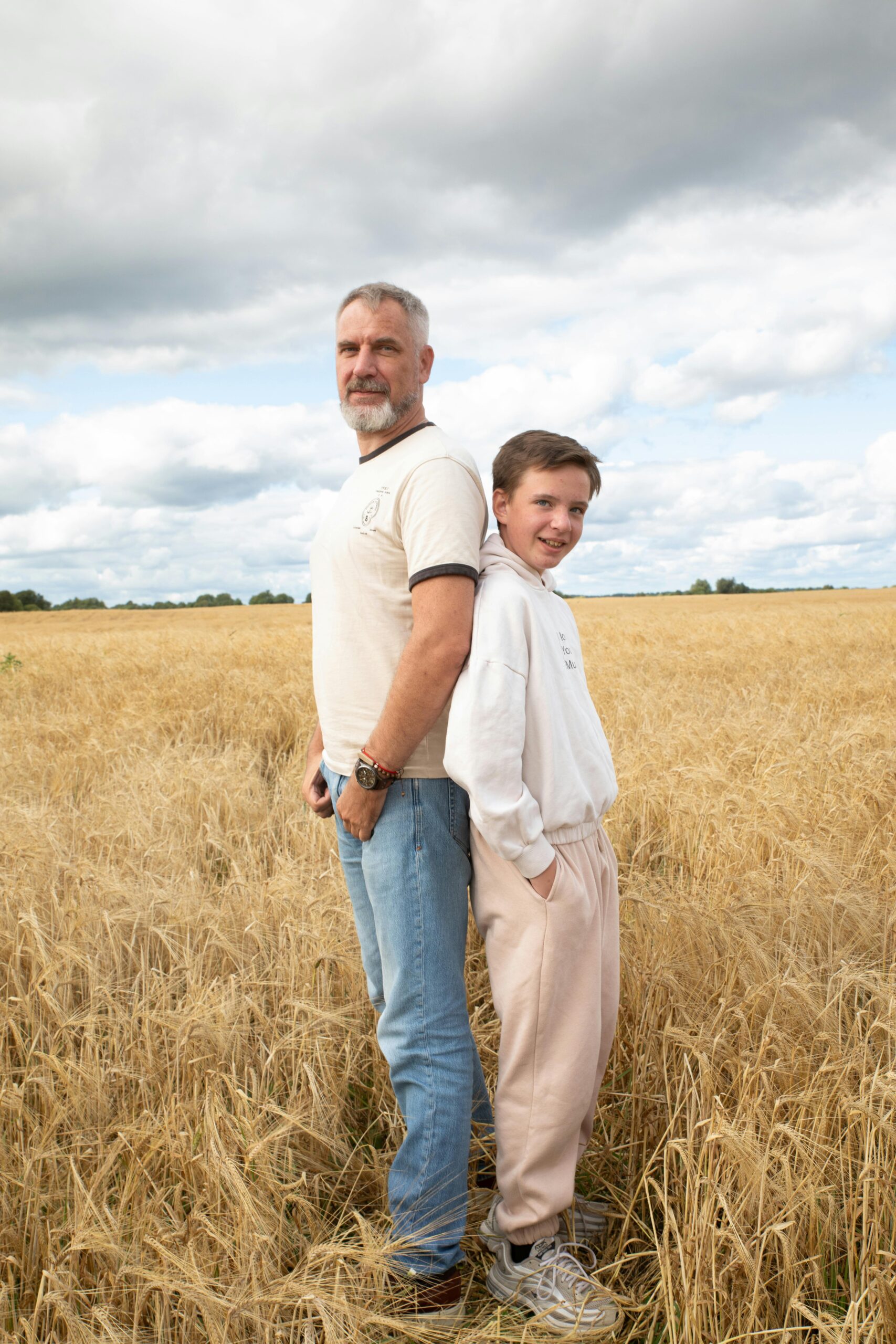 Father and son standing back-to-back in a golden wheat field under a cloudy sky.